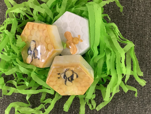 The image displays a collection of fun-shaped soaps floating on a green, grass-like surface. These soaps are crafted to resemble various playful shapes, including a bee and a hexagon.